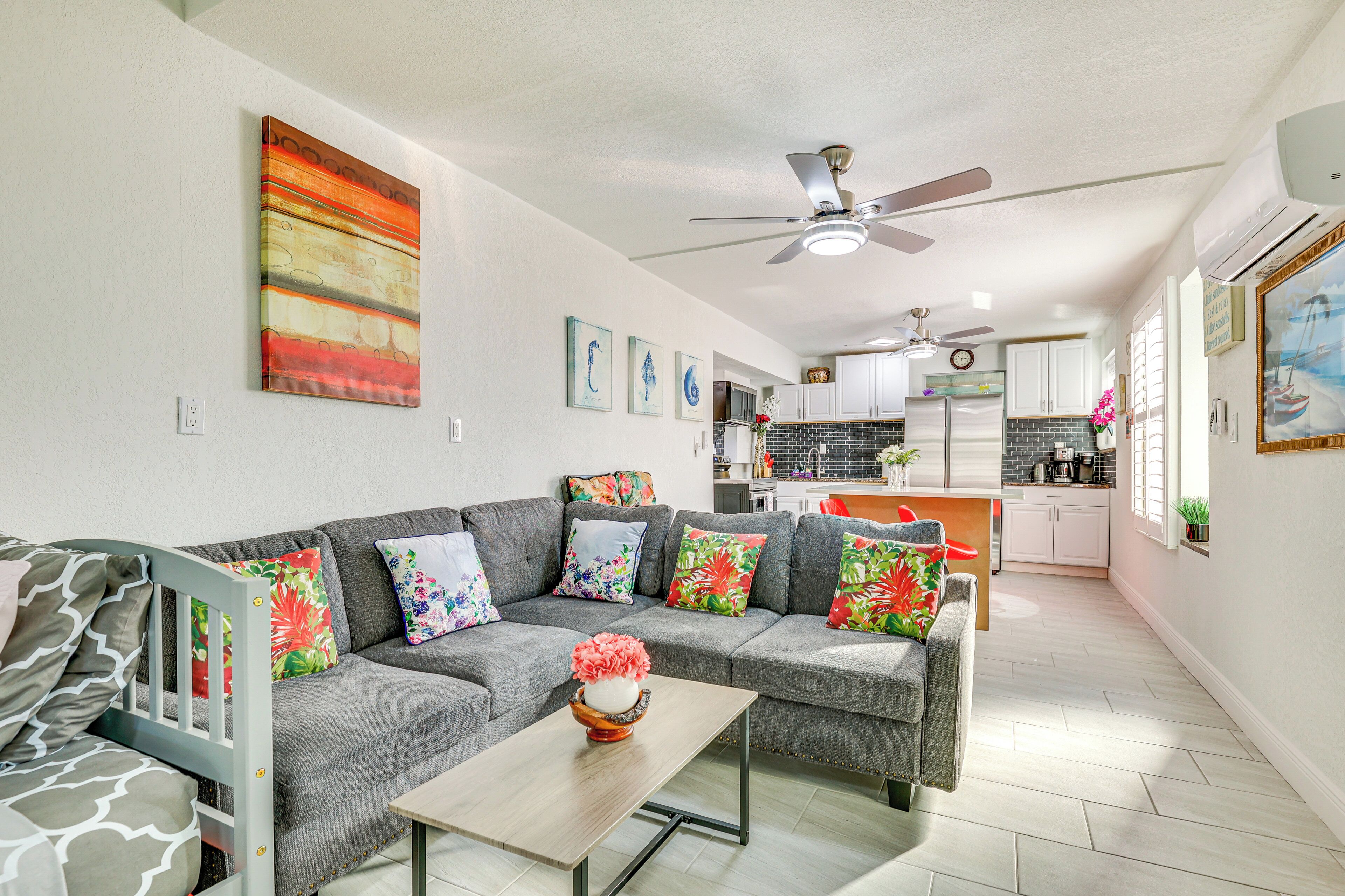 A modern living room with a gray sectional sofa, a wooden coffee table, and colorful pillows. A ceiling fan and air conditioning unit are visible above.