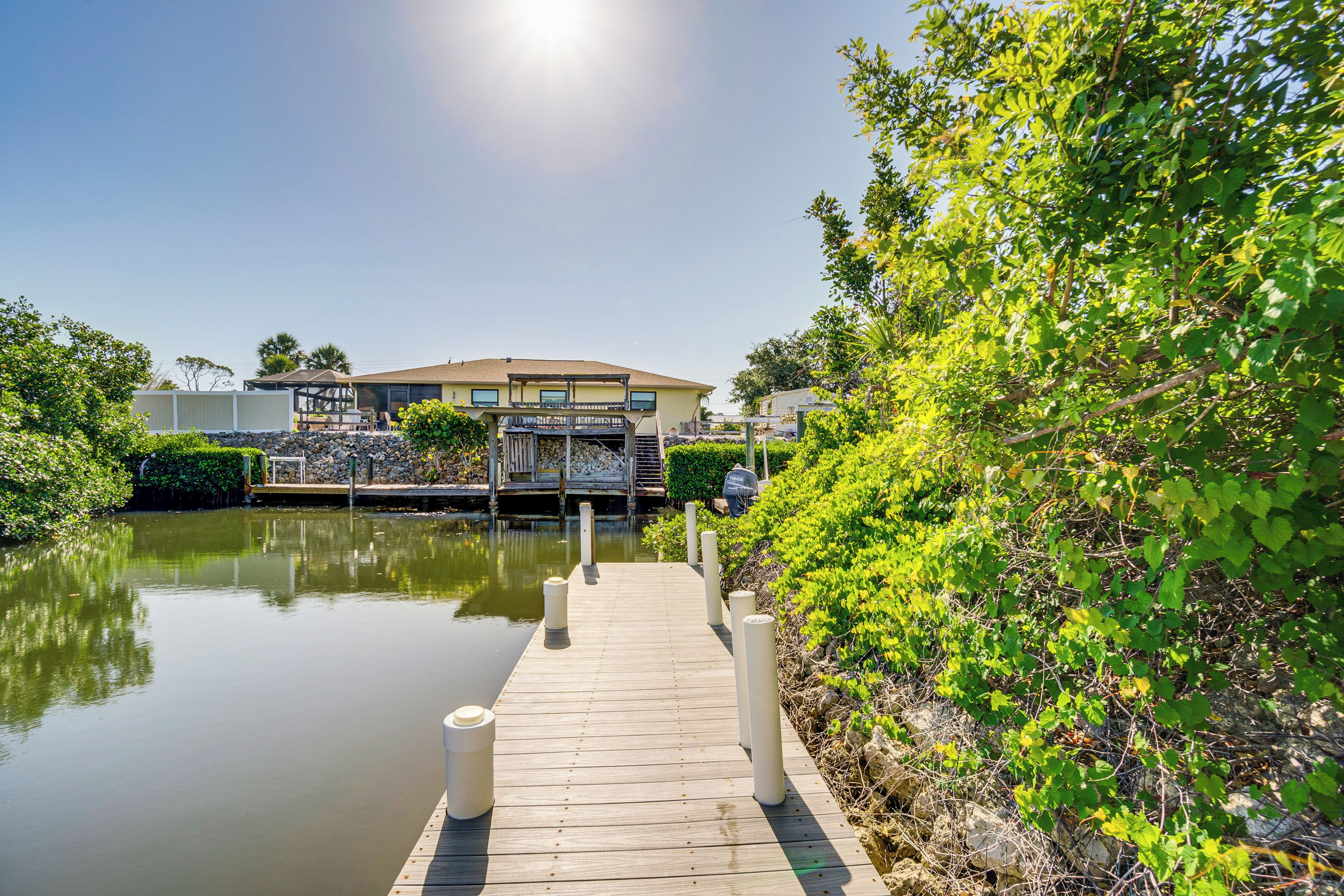A wooden dock leading to a house with a balcony, surrounded by greenery and a calm body of water.