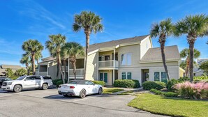 A multi-story residential building with a balcony on the second floor, a driveway with two white cars, and palm trees in the front yard.