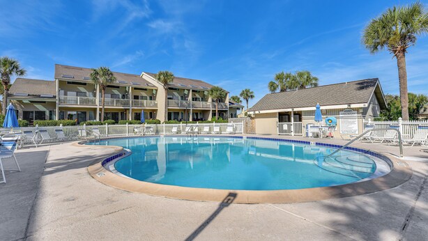 A swimming pool with lounge chairs and umbrellas, surrounded by a building with balconies and palm trees.