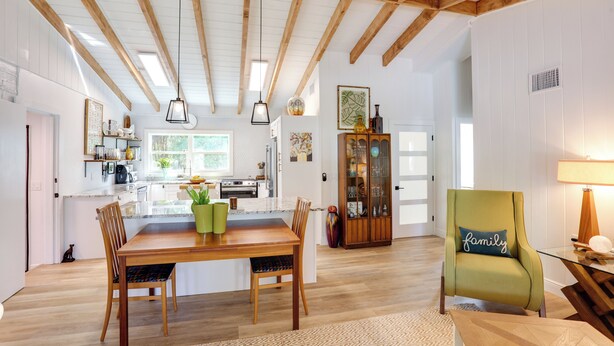 A spacious living area with a wooden dining table, chairs, and a green armchair. A kitchen is visible in the background with modern appliances and a window.