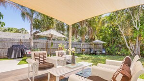 An outdoor patio with white furniture, including a sofa and chairs, under a canopy. There is a grill and a dining table with a vase and flowers.