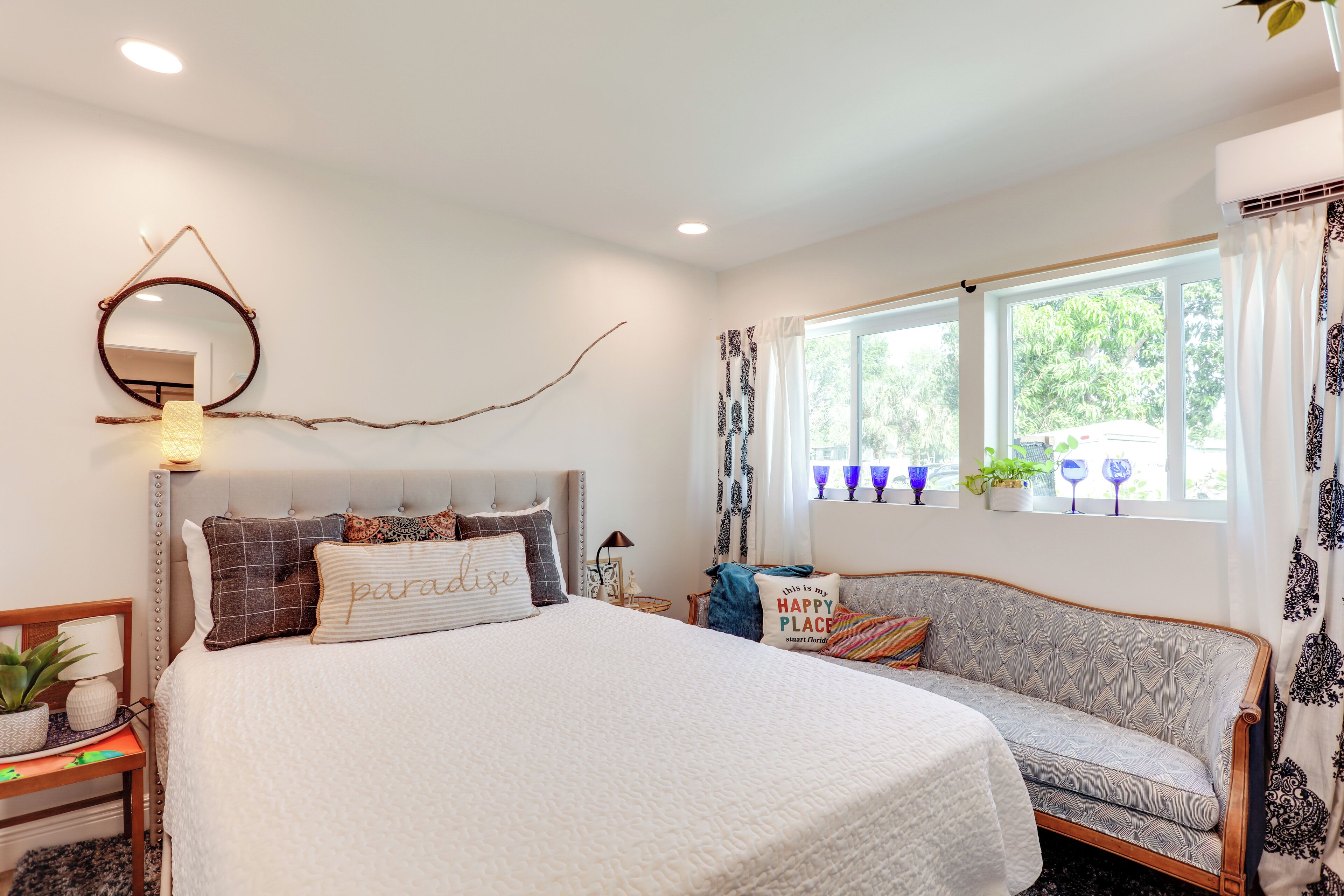 A bedroom with a bed, a sofa, a window with curtains, and a wall-mounted mirror.