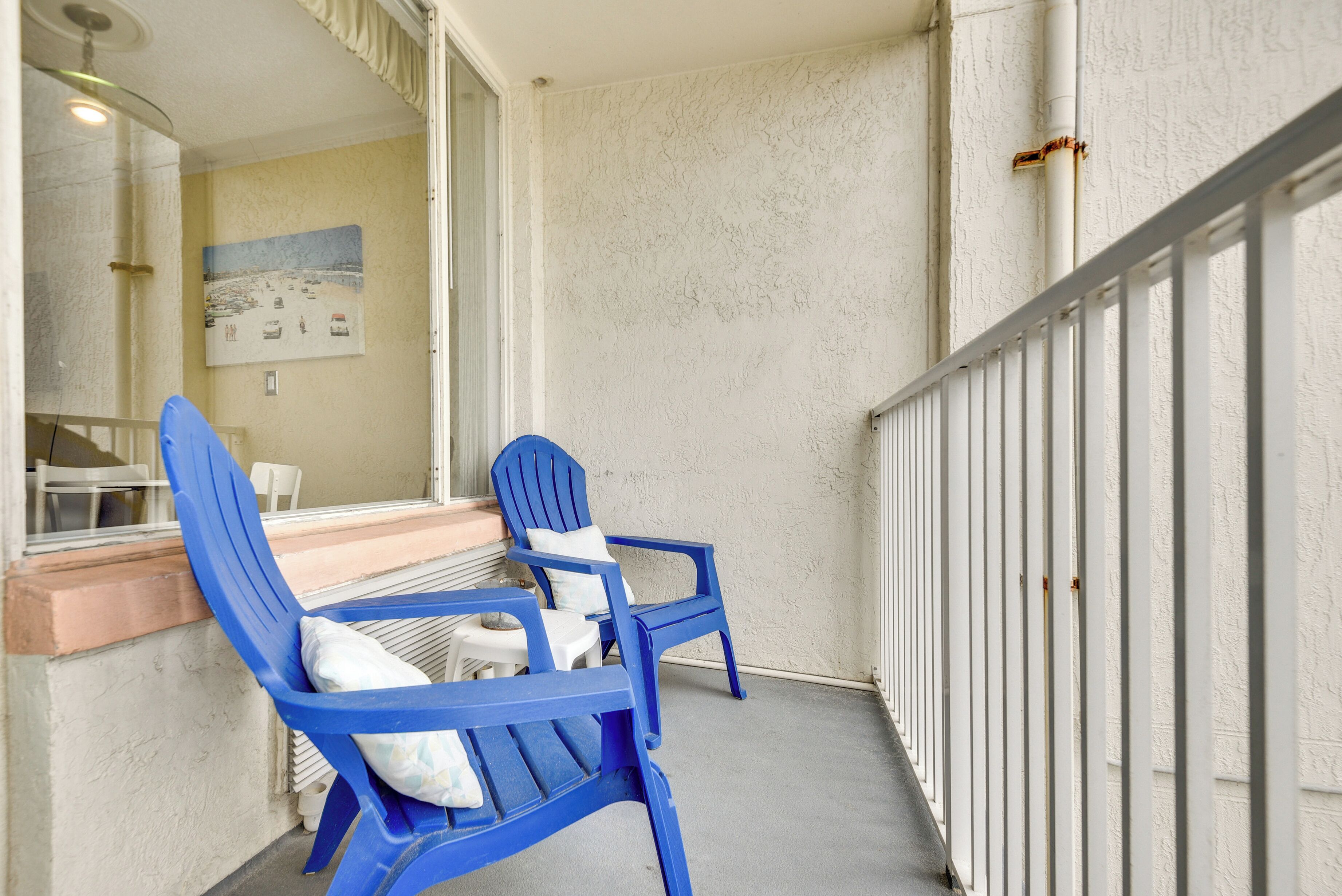 A balcony with two blue chairs and a small table, a white railing, and a painting on the wall.