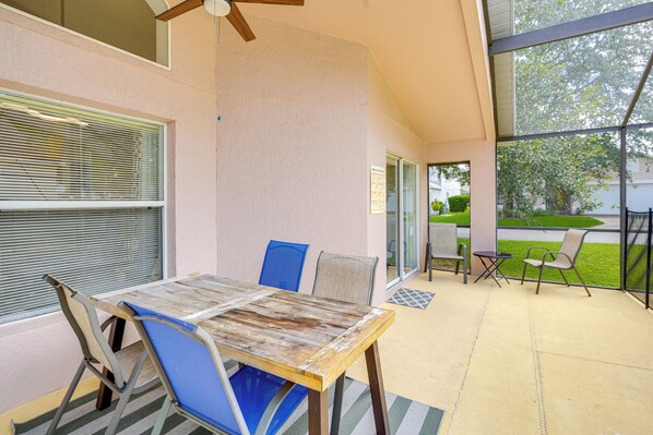 Un patio con una mesa y sillas de madera, un ventilador de techo y vistas a un césped verde y árboles.