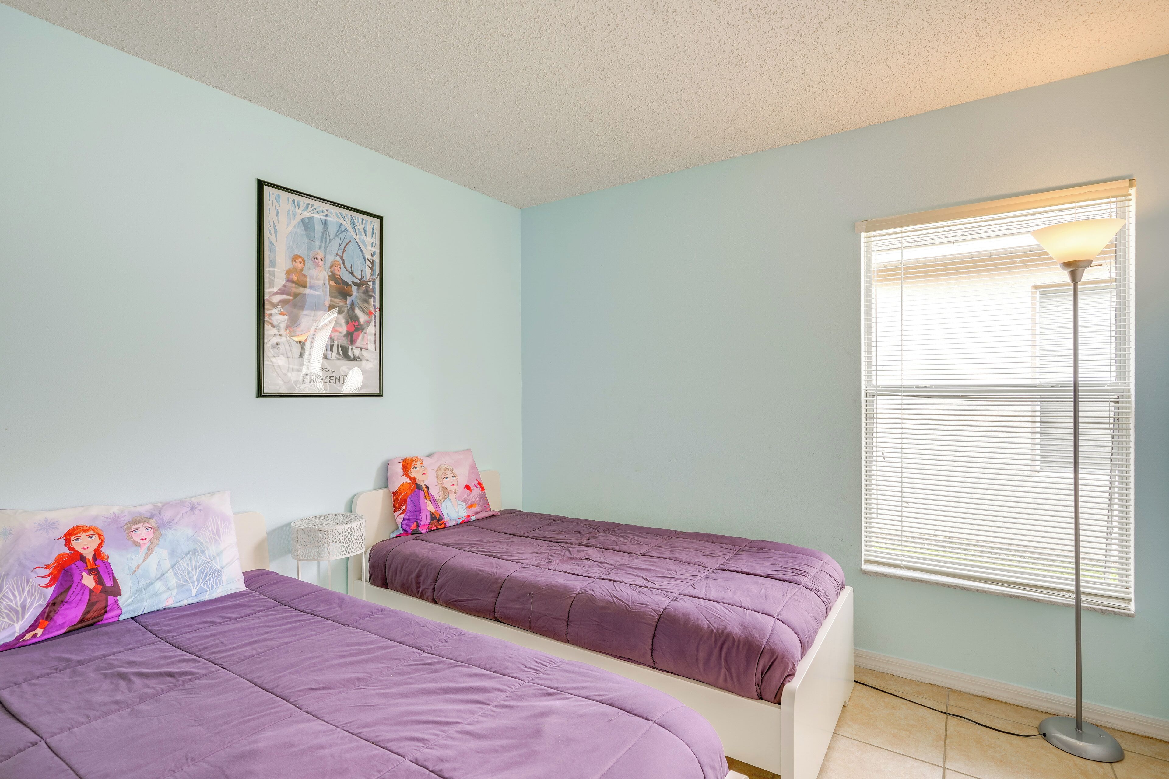 A bedroom with two beds, a window with blinds, a floor lamp, and a framed picture on the wall.
