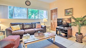 A living room with a leather sofa, a coffee table, a TV stand with a flat-screen TV, and a bookshelf. - Idyllic Apt w/ Pool Access - Walk to Beach! (Lauderdale-by-the-Sea)