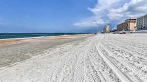 A sandy beach with tire tracks, a clear blue sky, and buildings in the background.