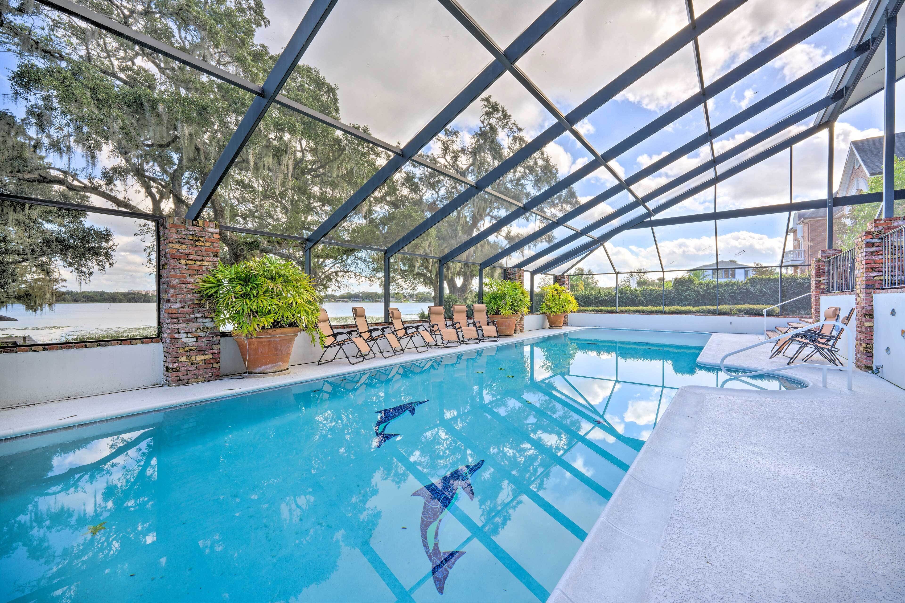 A swimming pool with clear blue water, surrounded by lounge chairs and potted plants, with a view of trees and a building in the background.