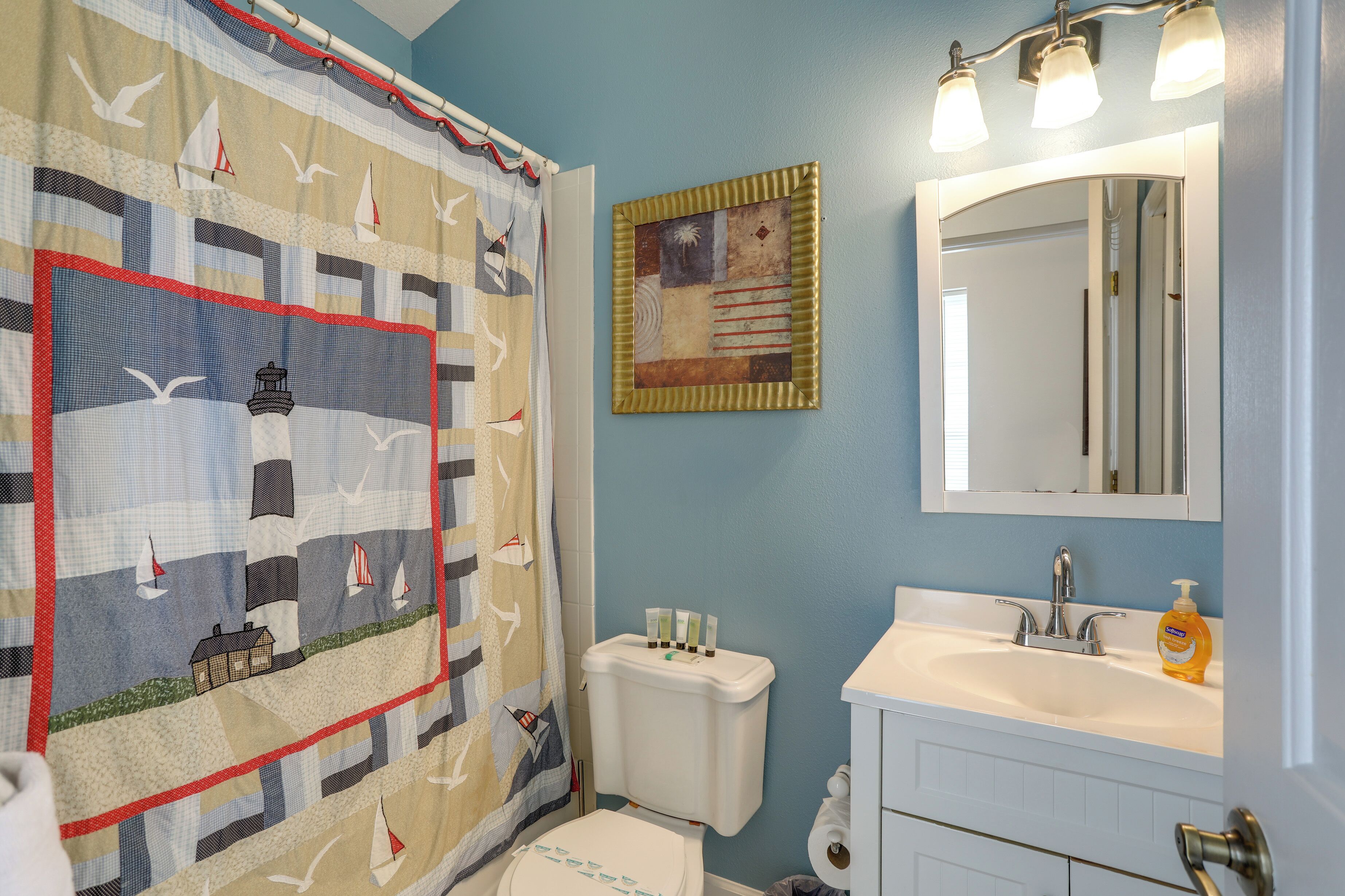 A bathroom with a quilted shower curtain, a toilet, a sink with a mirror, and a bottle of hand soap.
