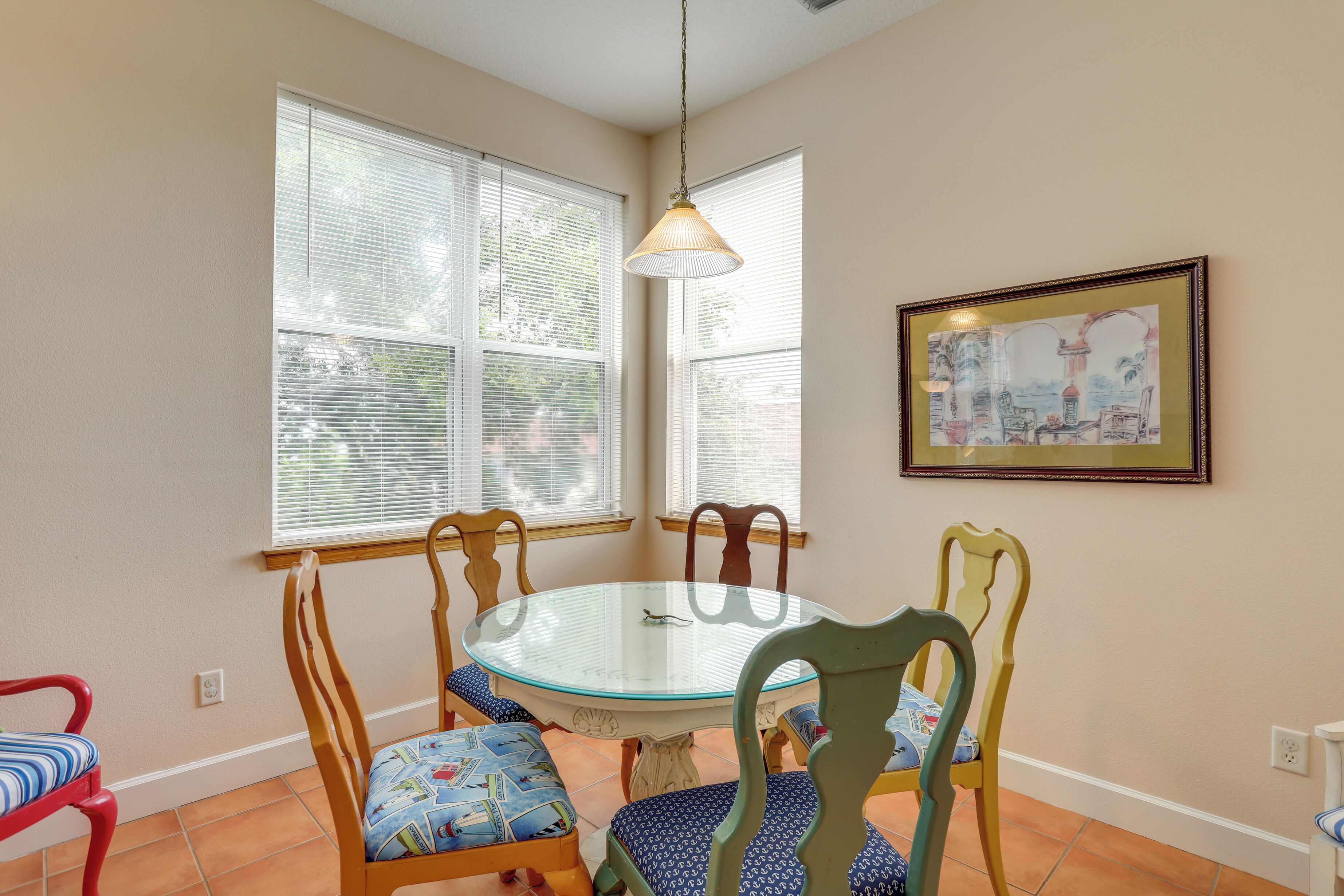 A dining area with a round glass table surrounded by wooden chairs with blue patterned cushions. There are two windows with blinds, a hanging light fixture, and a framed picture on the wall.