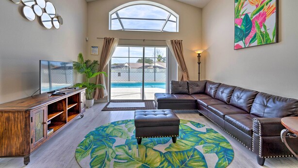 Ein Wohnzimmer mit einer Ledersofa, einem Fernseher, einem Holzkabinett und einem großen Fenster mit Blick auf einen Pool.