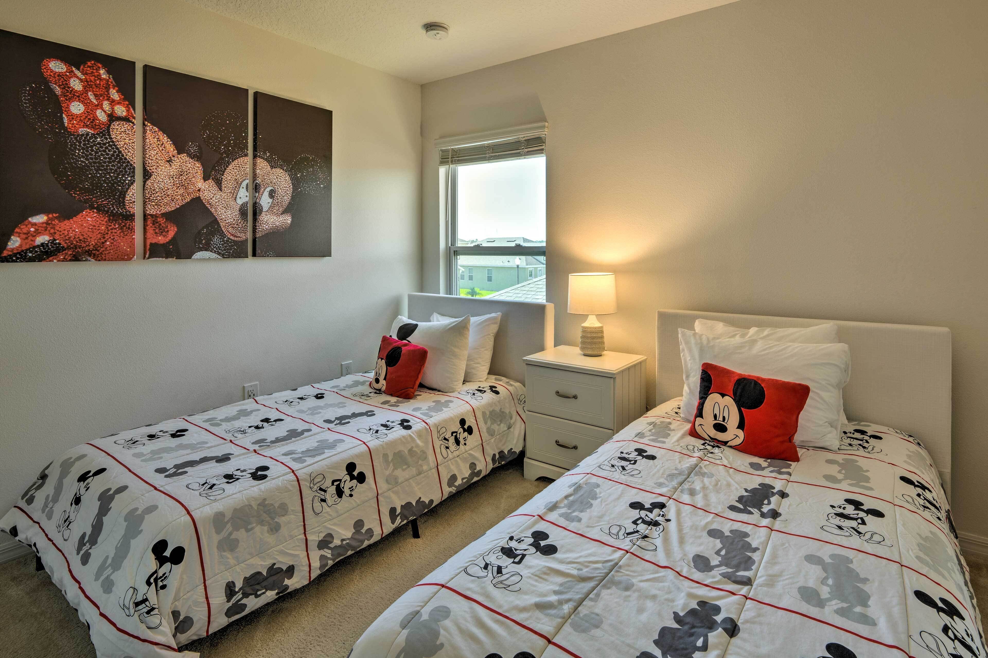 Ein Hotelzimmer mit zwei Betten, beide mit Bettwäsche im Mickey-Mouse-Design. Ein Fenster mit Jalousien, eine Nachttischlampe und ein Wandbild über den Betten.