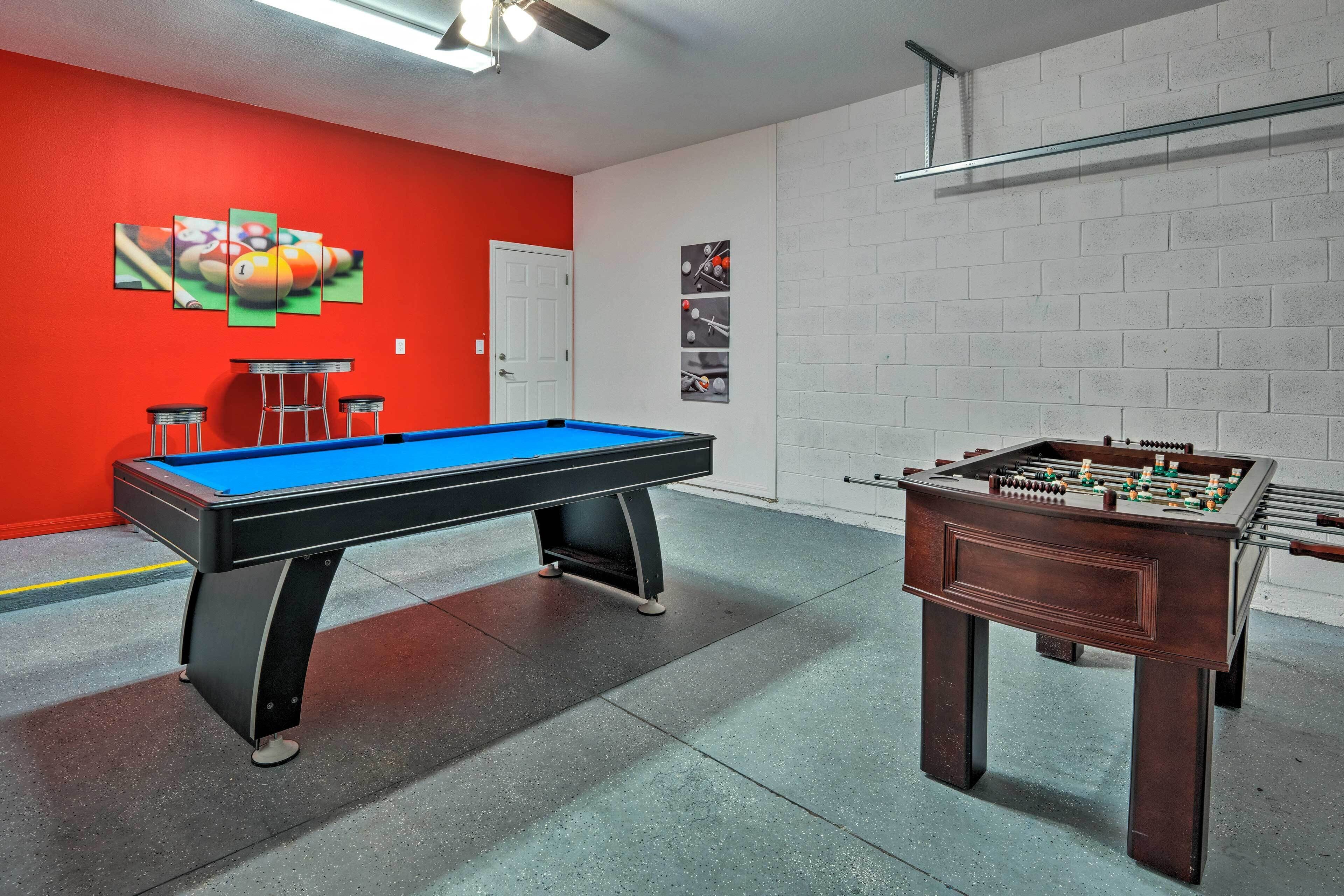A pool table and a foosball table in a room with a red wall and white brick wall.