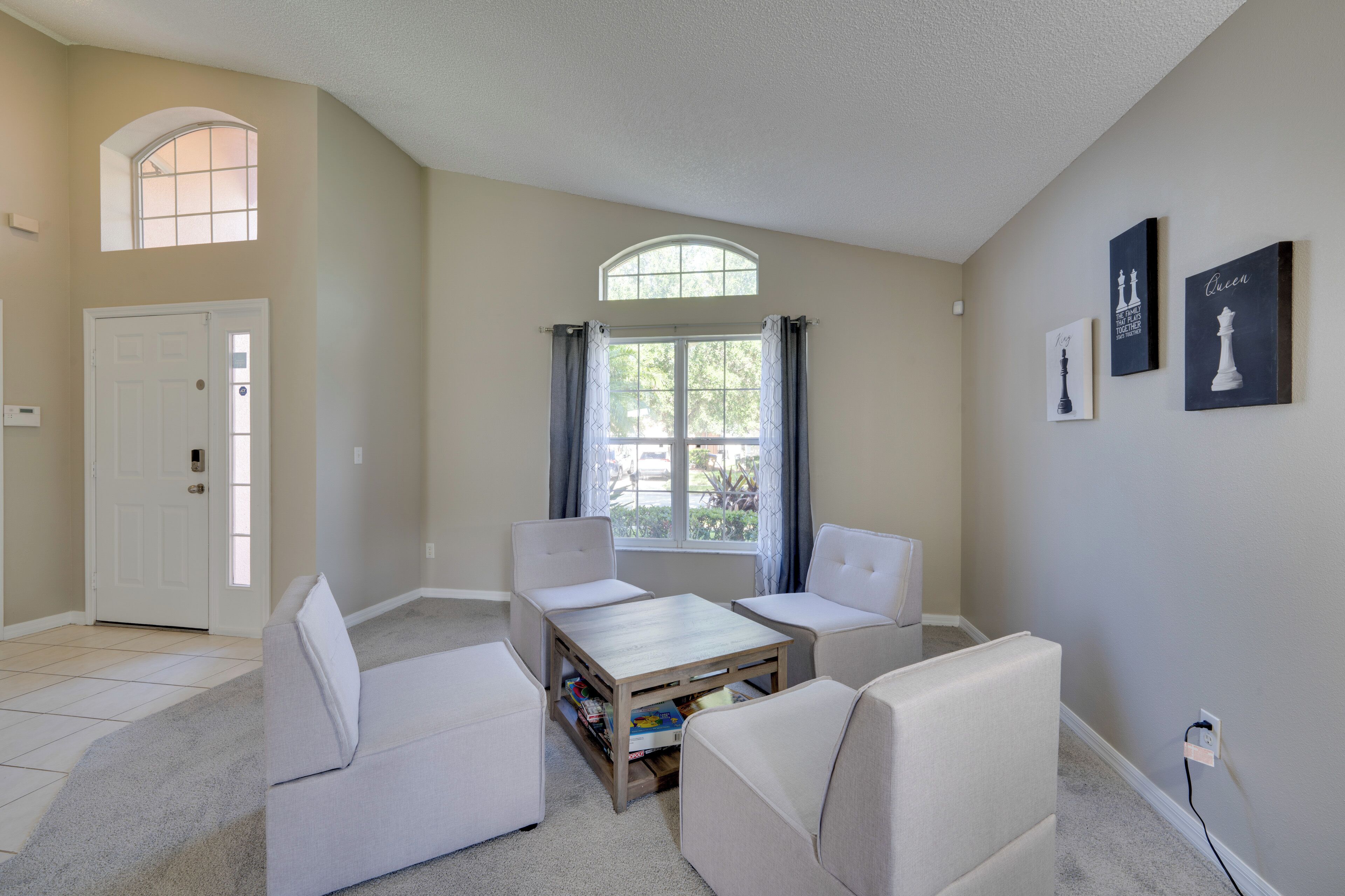 A living room with a high ceiling, a large window with curtains, a white door, and a small table with books.