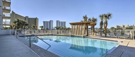A rooftop swimming pool with a pergola and lounge chairs, surrounded by palm trees and high-rise buildings.