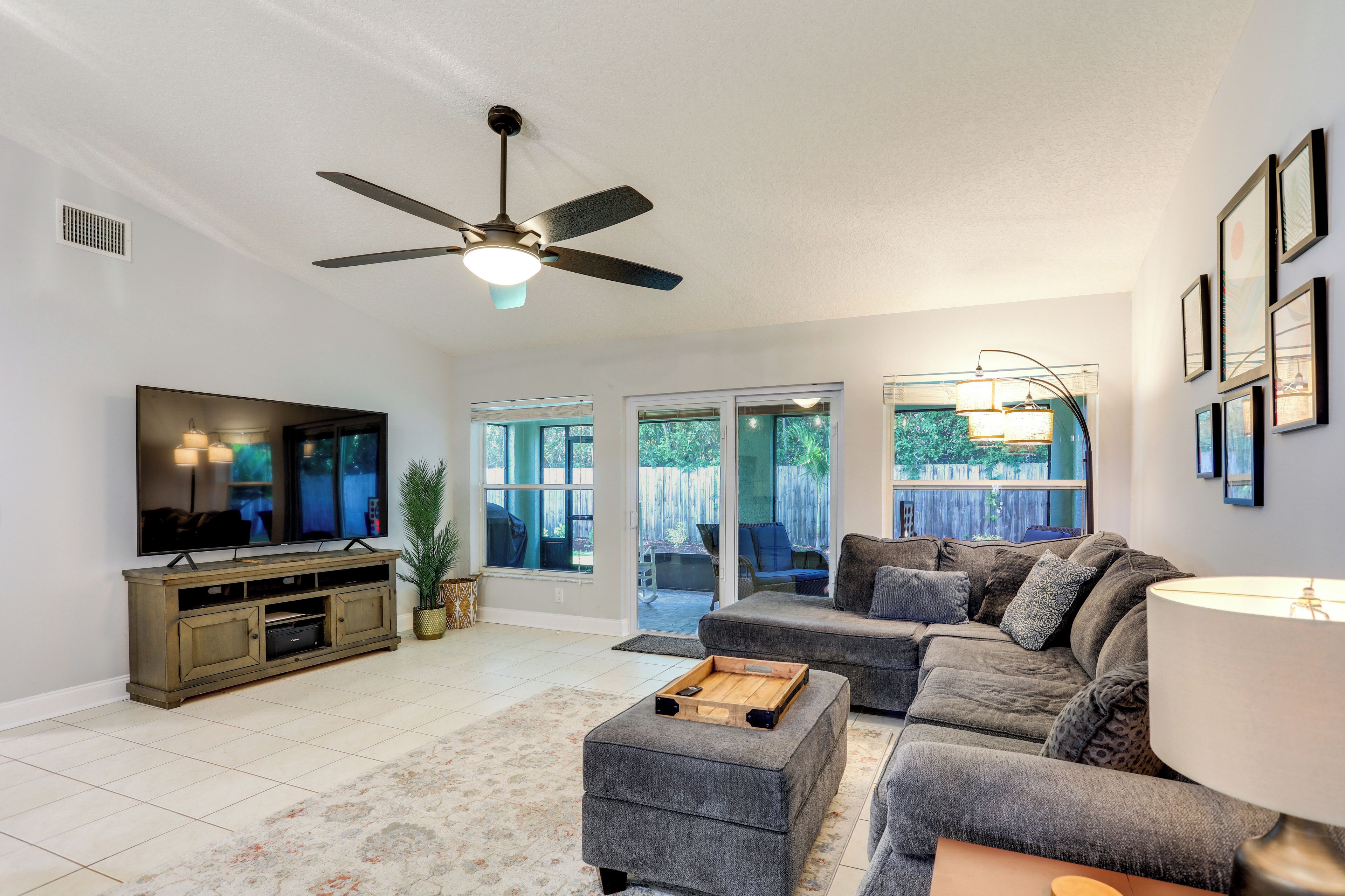 A living room with a flat-screen TV, grey sofas, a ceiling fan, and a large window.