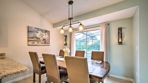 Un comedor con una mesa de madera y seis sillas, una araña, un cuadro y una ventana con cortinas.