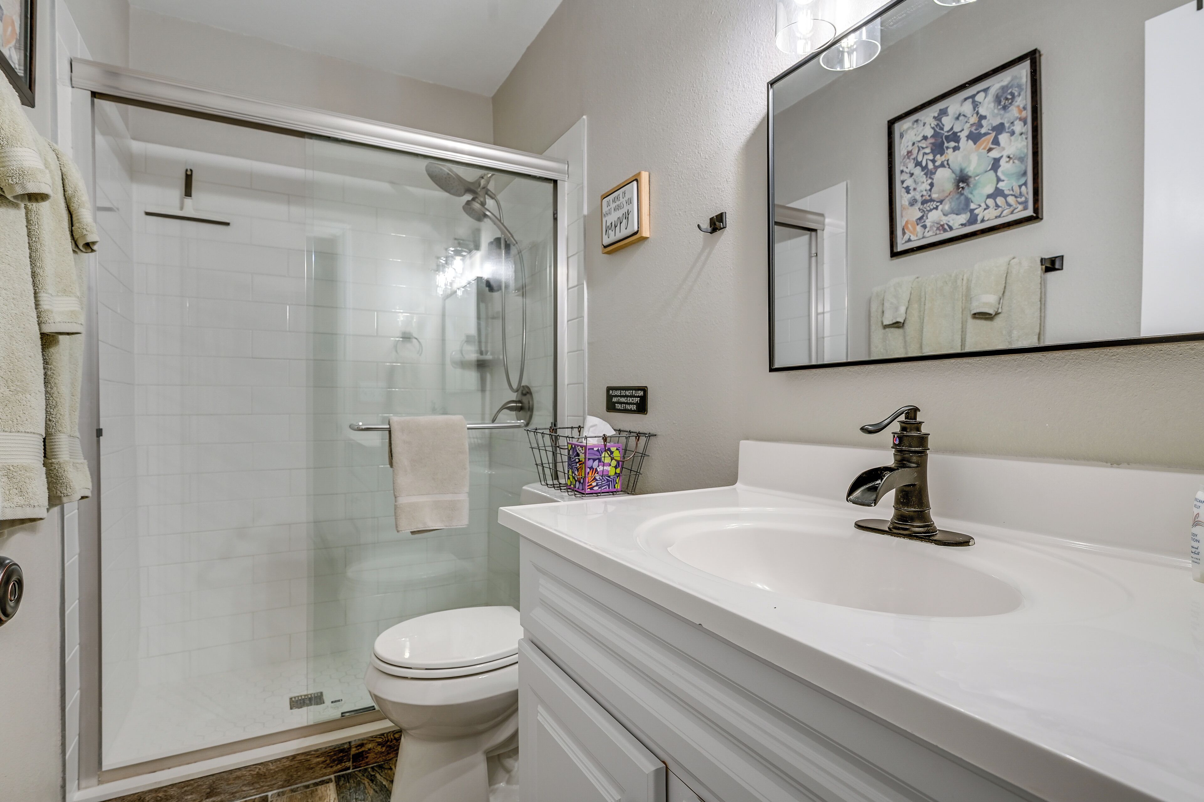 A bathroom with a glass-enclosed shower, a white sink, a toilet, and a mirror with a floral picture.