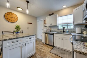 A kitchen with white cabinets, a stainless steel dishwasher, a clock, pendant lights, and a window with blinds.