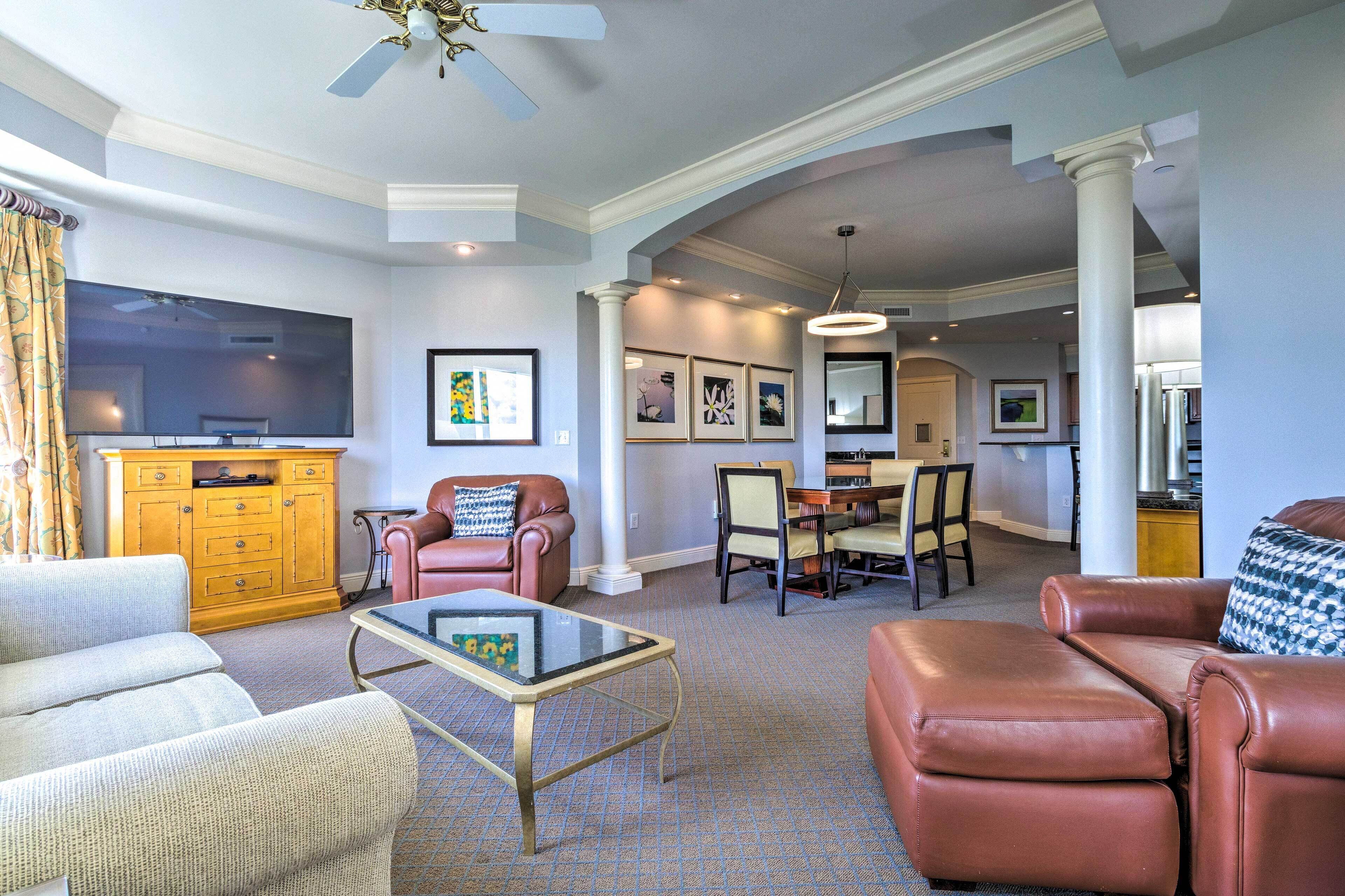 A spacious living room with a flat-screen TV, leather sofas, a dining area with chairs, and a kitchen in the background.