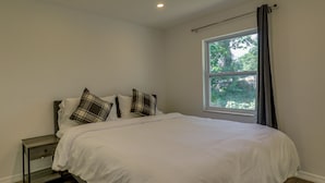 Quarto com cama, mesa de cabeceira, janela com cortinas e vista para o verde lá fora.
