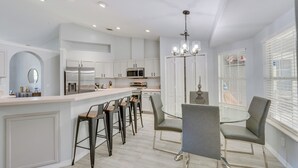 Une cuisine moderne avec des meubles blancs, un îlot central doté de tabourets, une table à manger avec des chaises et un lustre.