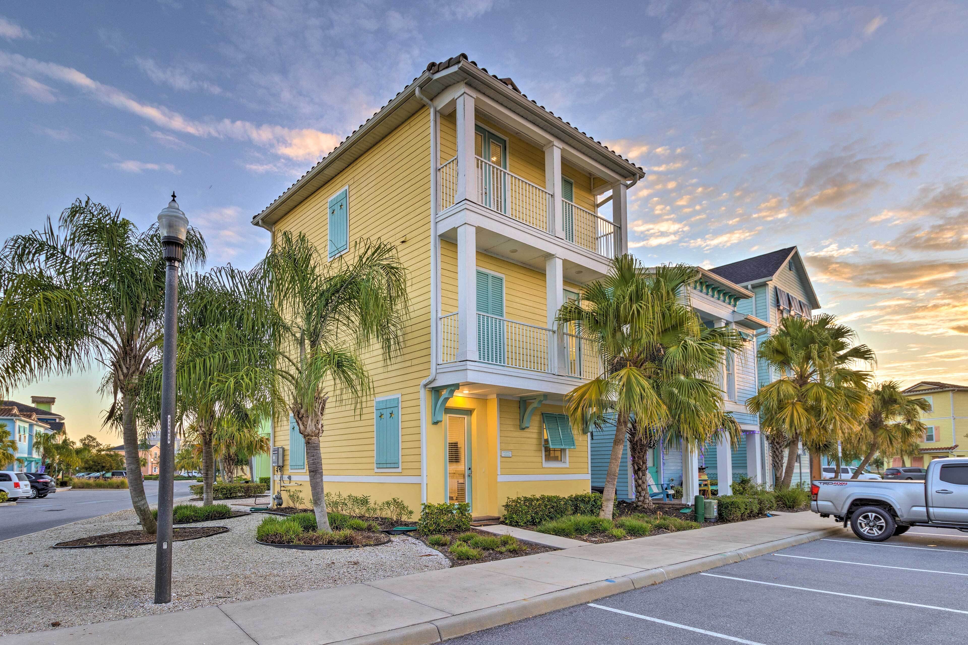 A two-story yellow building with blue shutters and a balcony, surrounded by palm trees and a parking lot.