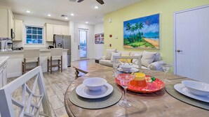 A modern kitchen with white cabinets, a dining area with a table set for two, and a living room with a couch and a painting of a beach scene. - 3 Mi to Disney: Home w/ Hot Tub in Margaritaville! (Kissimmee)