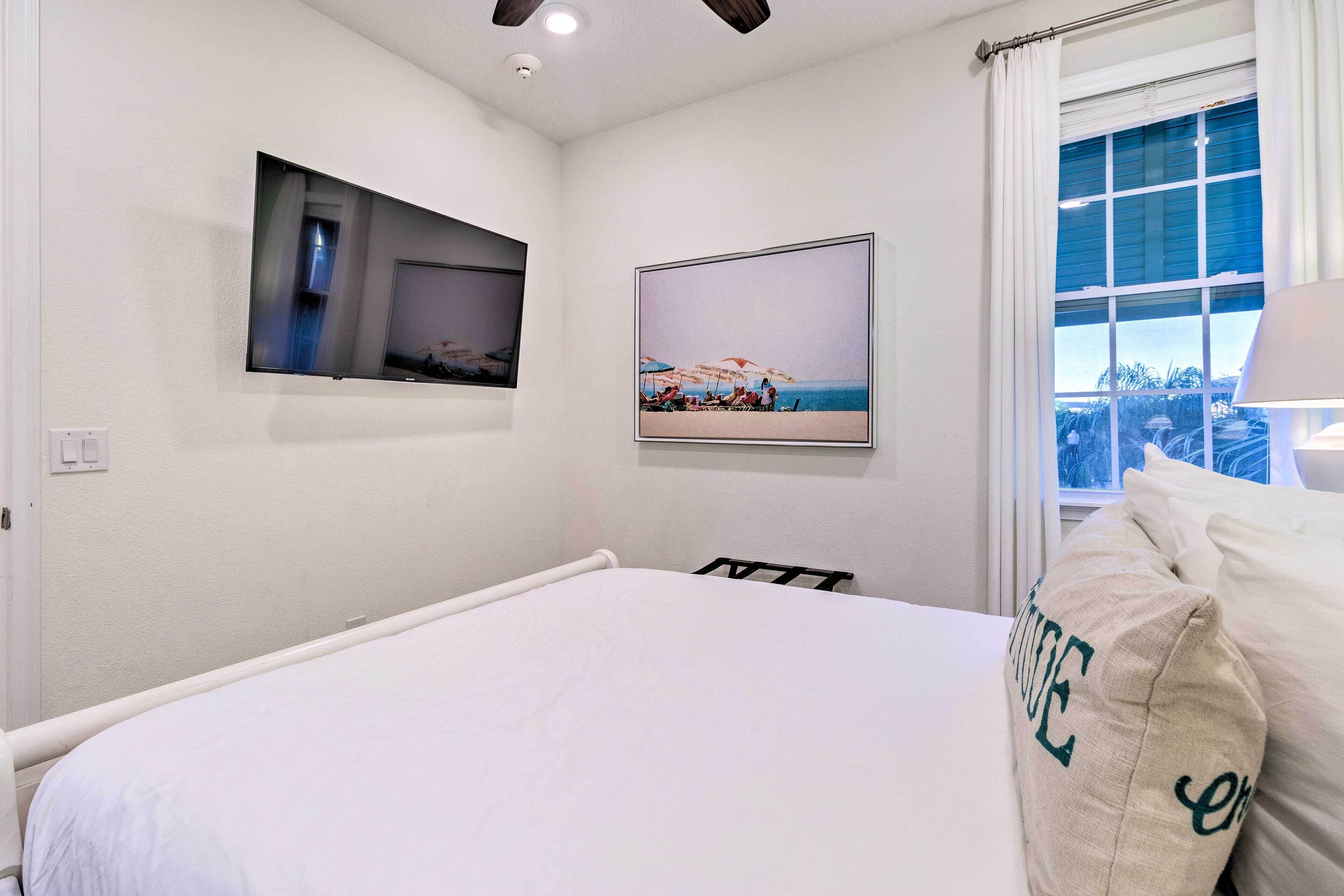 A bedroom with a bed, a wall-mounted TV, a window with curtains, and a beach-themed painting.