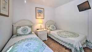 A bedroom with two single beds, a bedside table with a lamp, a framed picture on the wall, and a TV mounted on the wall. - Pool + Lanai: Emerald Isle Resort Home! (Kissimmee)