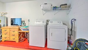 A laundry area with two white washing machines, a wooden dresser with drawers, a shelf with laundry baskets, and a television. - Pool + Lanai: Emerald Isle Resort Home! (Kissimmee)