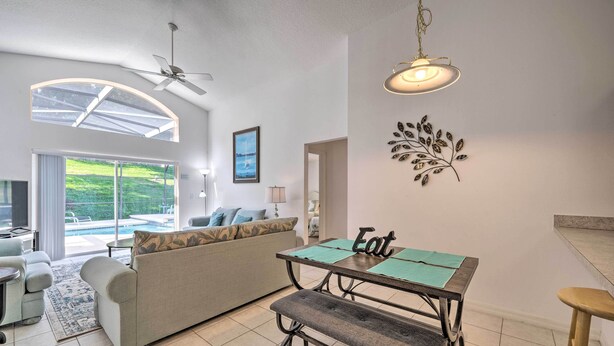 A modern living room with a sofa, a dining table with a bench, a ceiling fan, and a view of a pool through large windows. - Pool + Lanai: Emerald Isle Resort Home! (Kissimmee)
