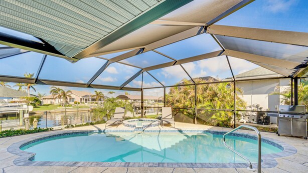 An outdoor swimming pool with a hot tub, surrounded by a covered patio area with a view of palm trees and houses. - Luxe Waterfront Oasis w/ Dock, Heated Pool & Spa! (Port Charlotte)