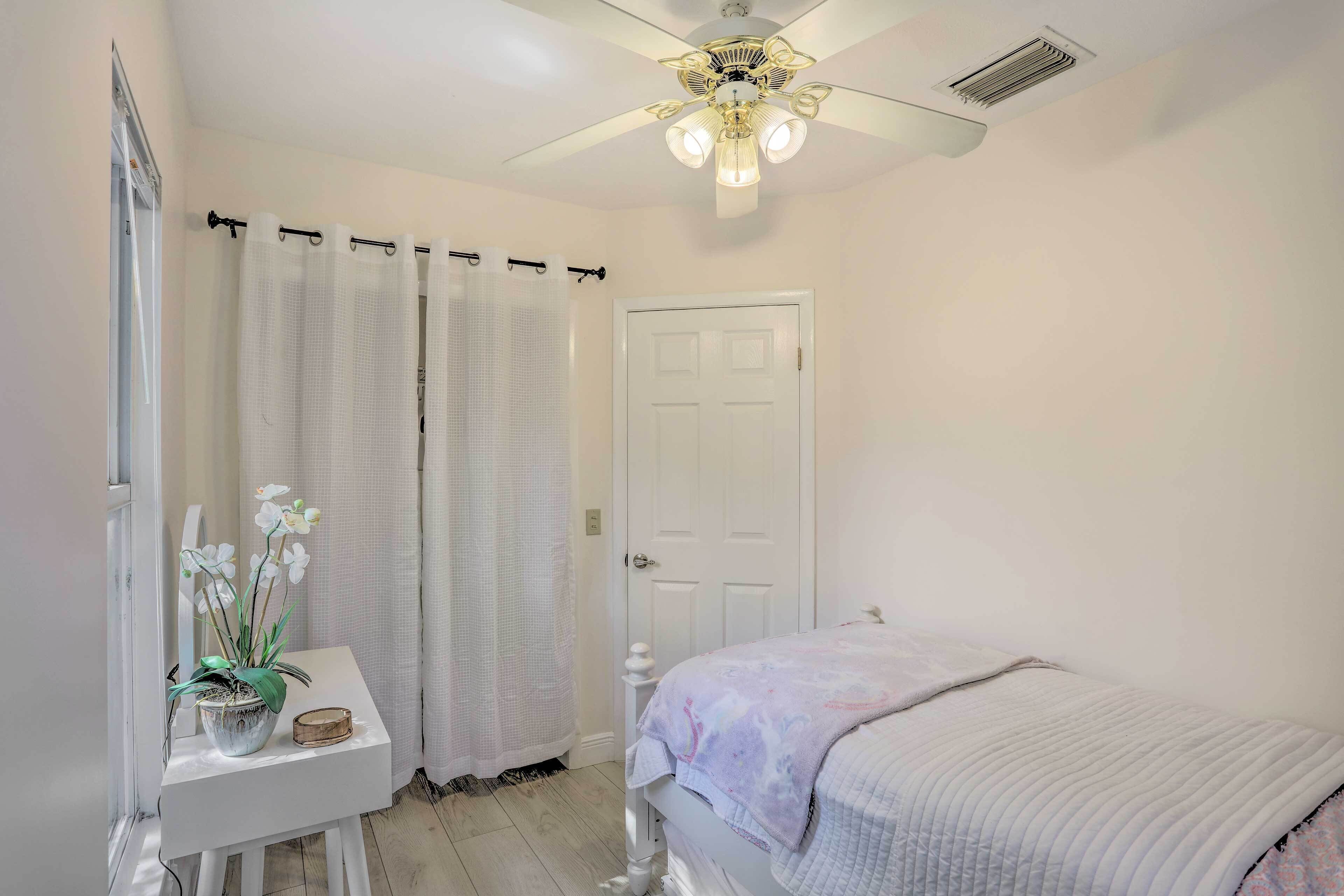 A bedroom with a bed, a ceiling fan, a door, and a small table with a plant.