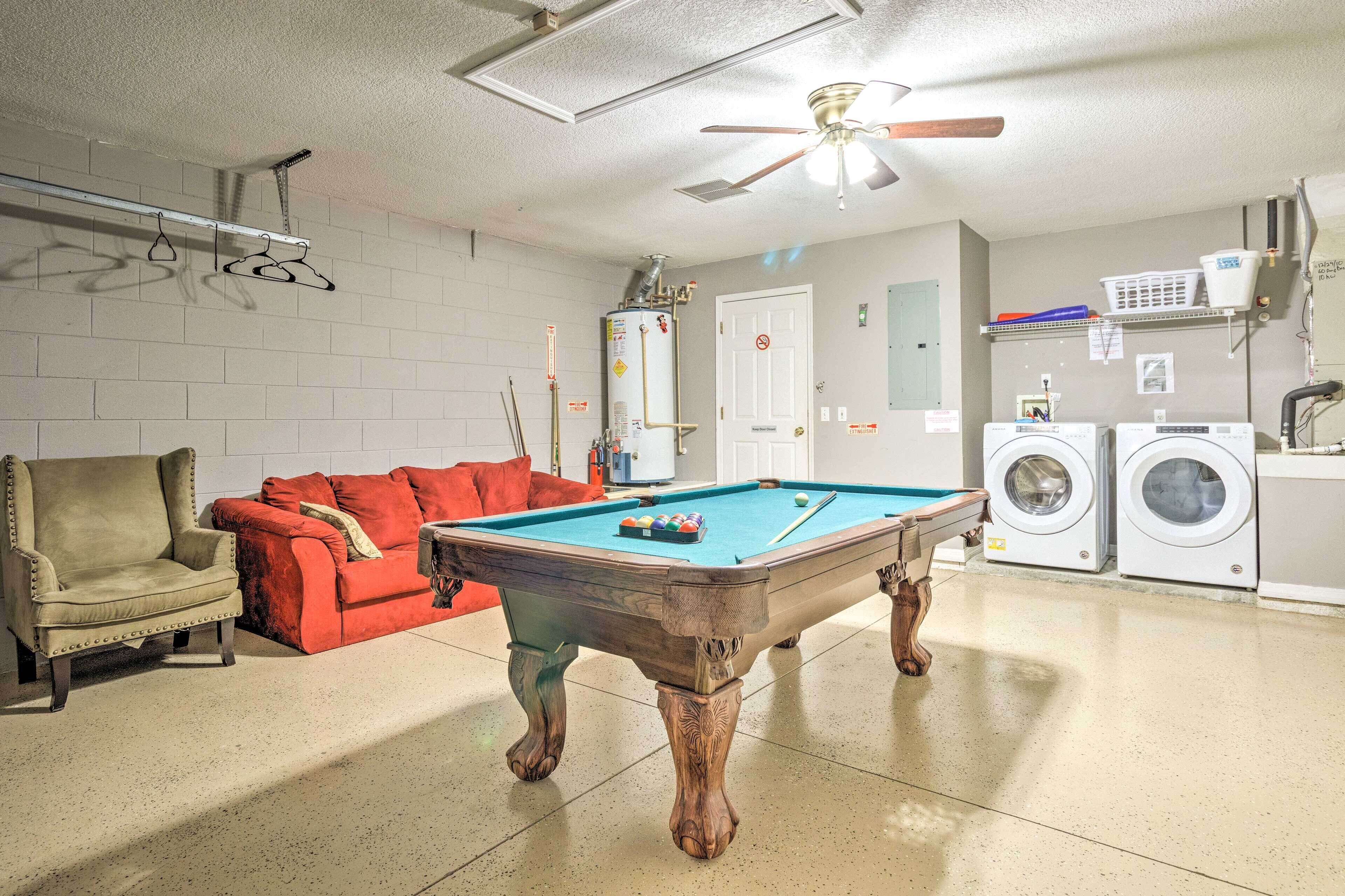 A pool table in a room with a ceiling fan, a red couch, a beige armchair, and a washing machine.
