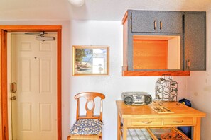 Um quarto com uma porta branca, uma cadeira de madeira com almofada estampada, uma mesa de madeira com um rádio e um porta-retrato, e um armário com organizador de metal.