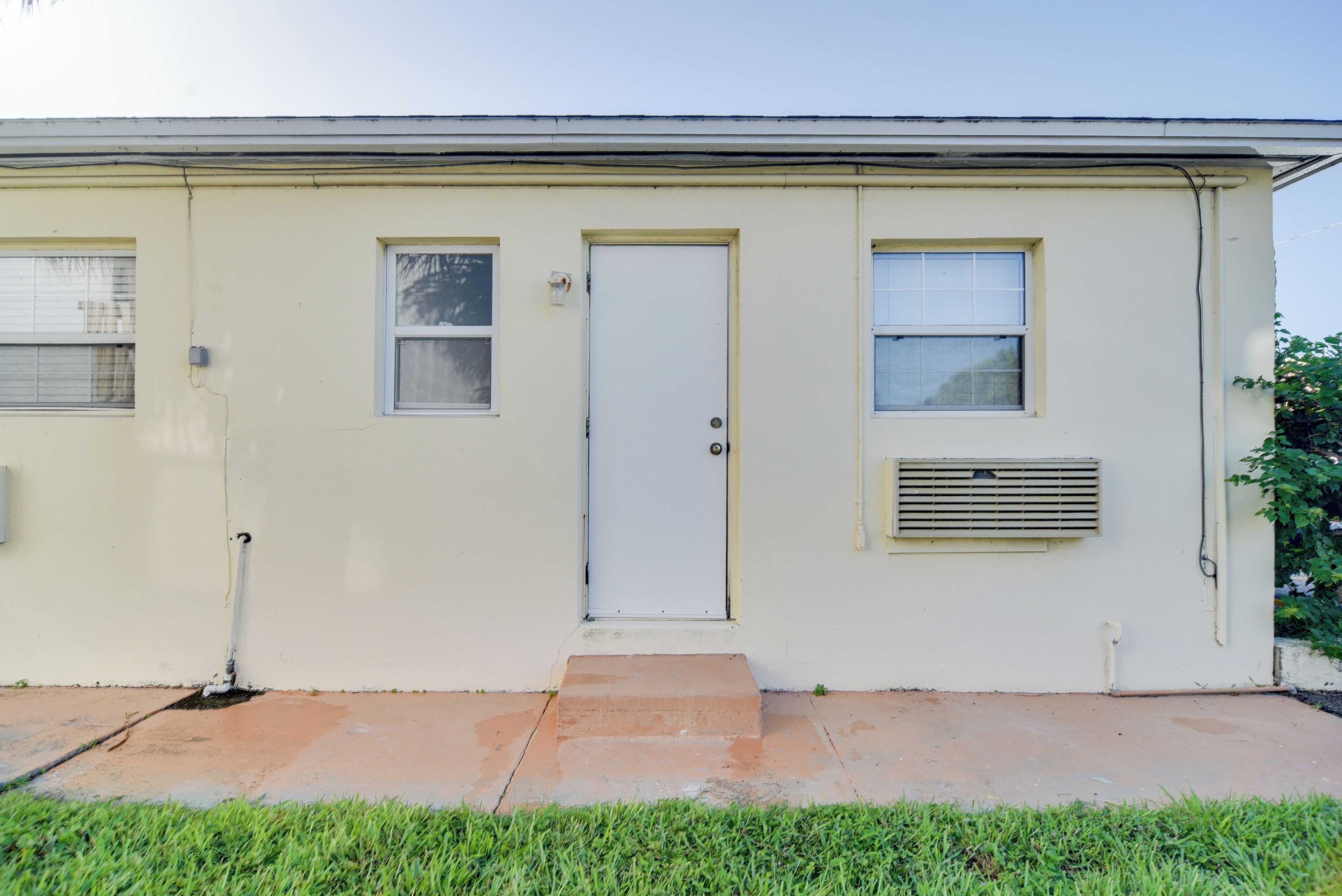 A single-story building with a white exterior, a front door, two windows, and a small air conditioning unit mounted on the wall.