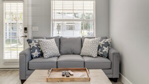 A grey sectional sofa with star and wave-patterned pillows, a wooden tray with remotes, and a white door with blinds. - Walk to Gulf: Madeira Beach Condo w/ Patio (Madeira Beach)