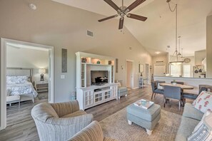 A spacious living room with a ceiling fan, a television on a white entertainment unit, a dining area with a round table and chairs, and a kitchen area visible in the background.