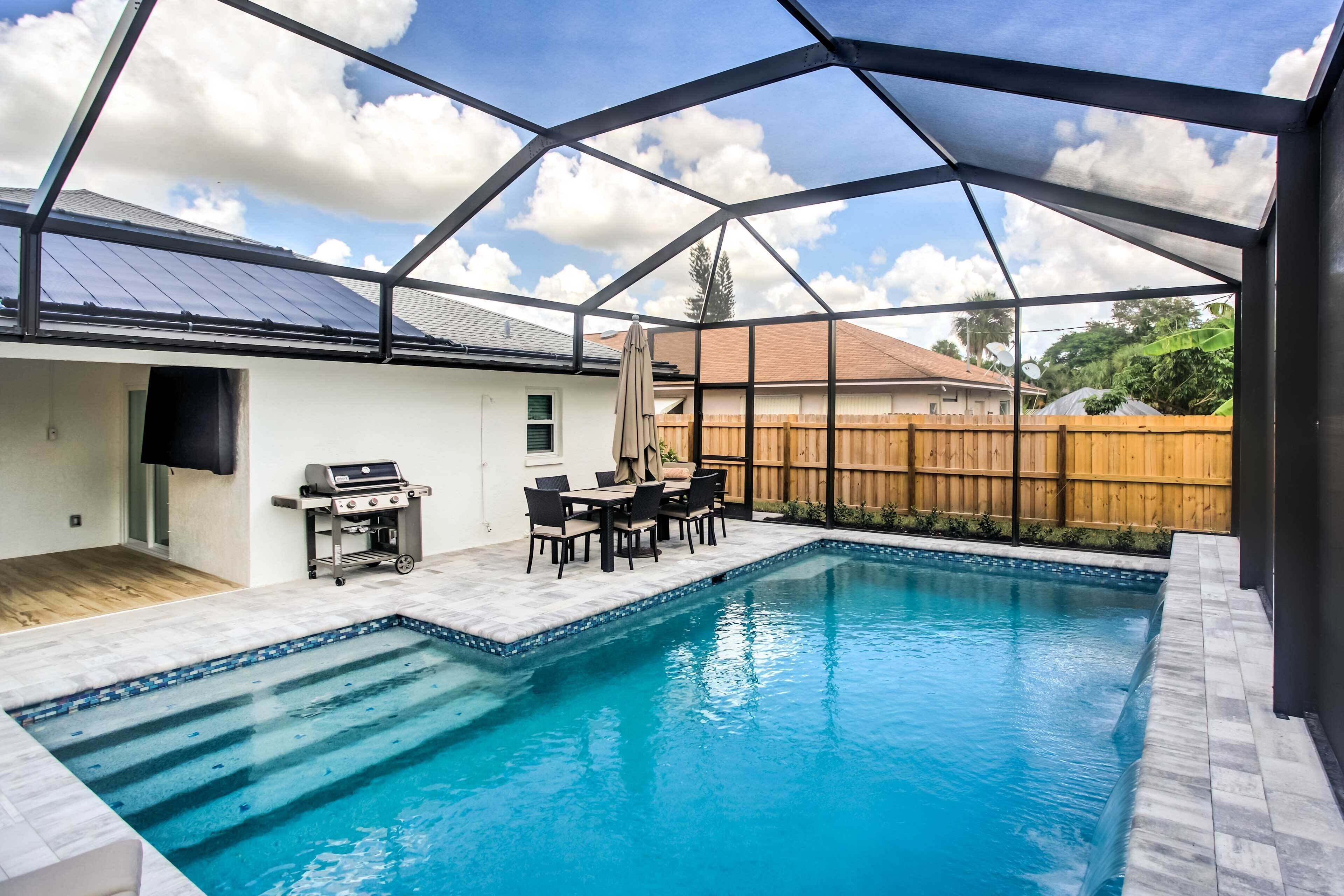 A covered outdoor swimming pool with a patio area, a grill, and a wooden fence in the background.
