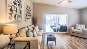 A living room with a sofa, coffee table, and a ceiling fan. There are abstract paintings on the wall and a view through a glass door to an outdoor area. - Naples Home w/ Lanai & Pool Near Vanderbilt Beach! (Naples)