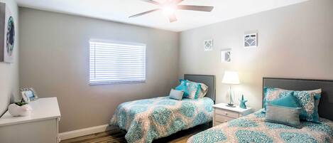 Une chambre avec deux lits, un ventilateur de plafond, une fenêtre avec des stores, une table de chevet avec une lampe et une commode.
