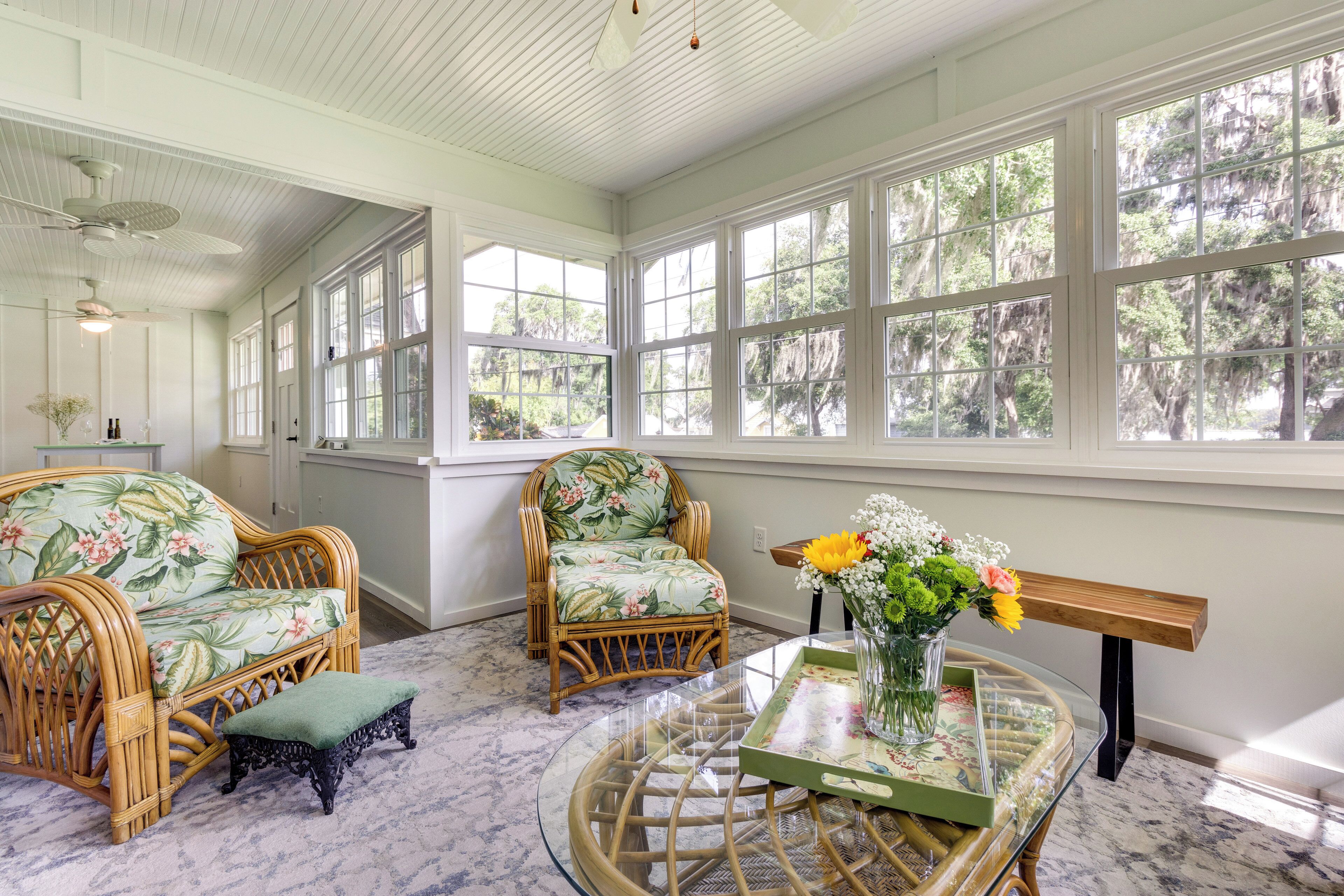A room with wicker furniture, large windows, and a glass table with a vase of flowers.