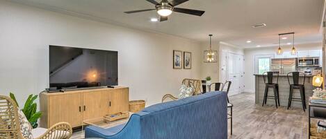 A modern living room with a TV, sofa, and chairs. There is a kitchen area with a bar and stools.