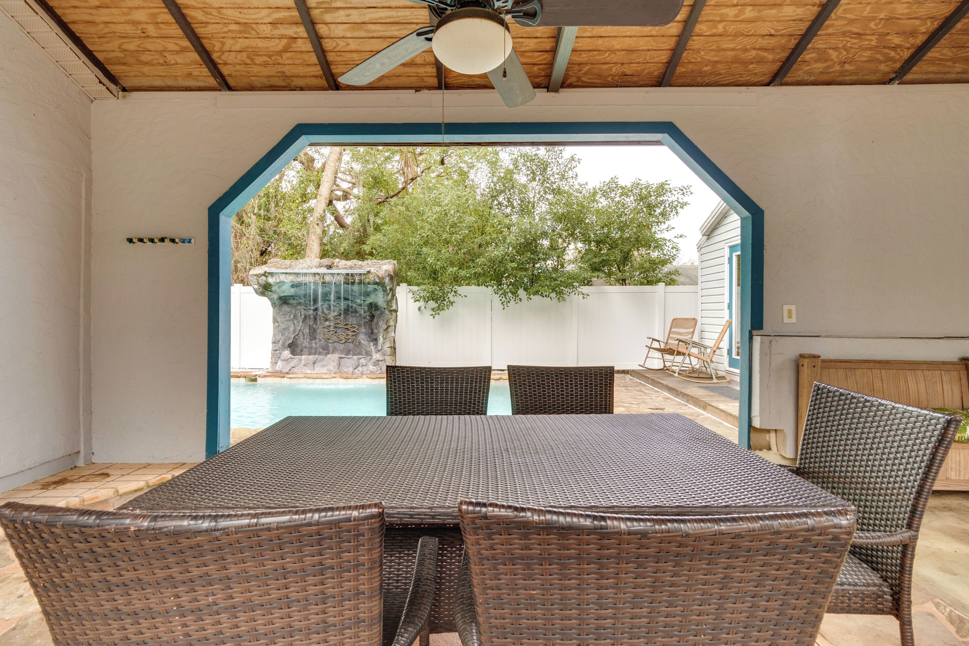 An outdoor patio with a table and chairs, a ceiling fan, and a view of a backyard with a swimming pool and a waterfall.