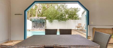 An outdoor patio with a table and chairs, a ceiling fan, and a view of a backyard with a swimming pool and a waterfall.