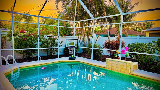 Zwembad met basketbalring, omgeven door groen en een hek.
