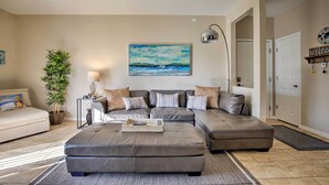A living room with a sectional sofa, a side table with a lamp, a painting of a seascape, and a welcome sign.