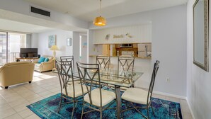 A modern living room with a glass dining table, beige sofas, and a kitchen area with white cabinets. - Dolphin & Manatee Viewing From 15th Floor & Pool! (Clearwater Beach)
